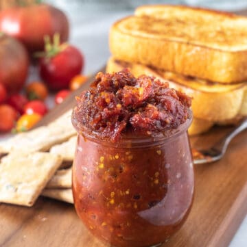 Close up of jar of tomato jam on wood platter with bread and tomatoes in background