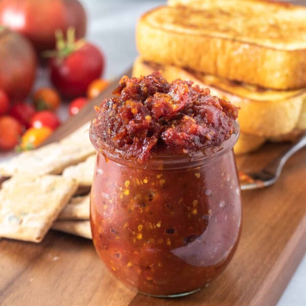 Close up of tomato jam in a jar on a wooden platter with accompaniments in the background
