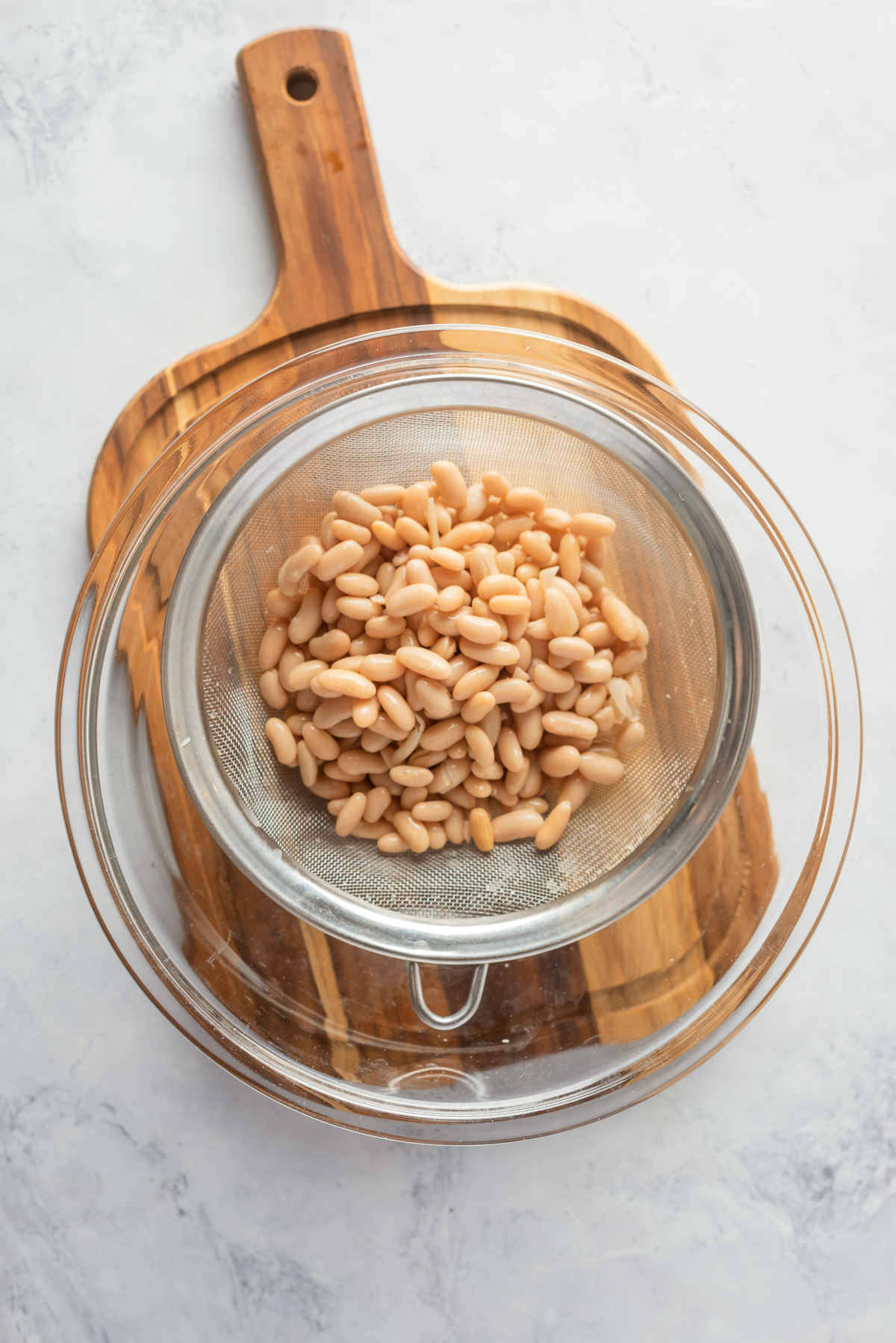Overhead view of drained cannellini beans on a strainer placed in a glass bowl on top of a wooden chopping board. 