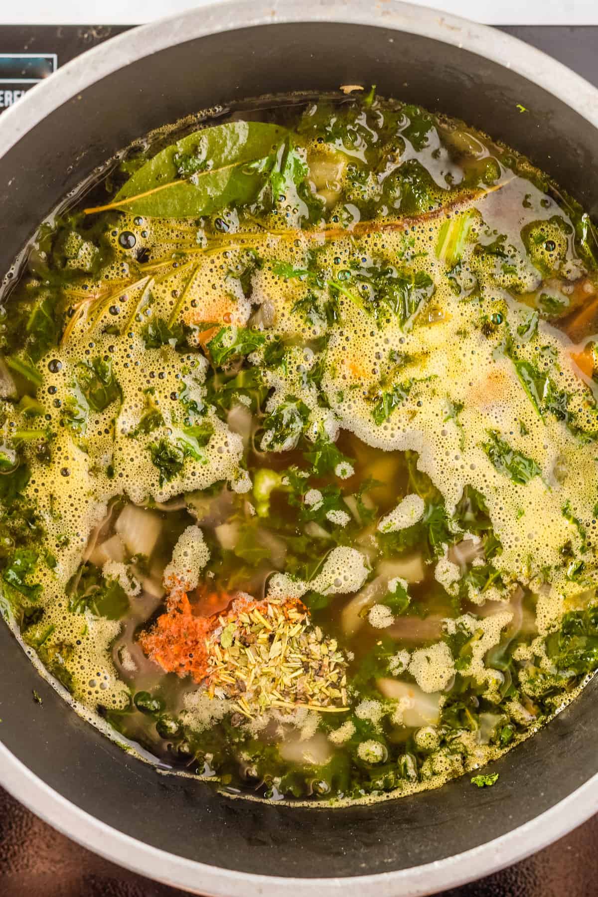 Overhead view of a large pot with boiled shallots, carrots, celery, and chard stalks, garlic, and bouquet garni added with salt, dried oregano and red pepper flakes.