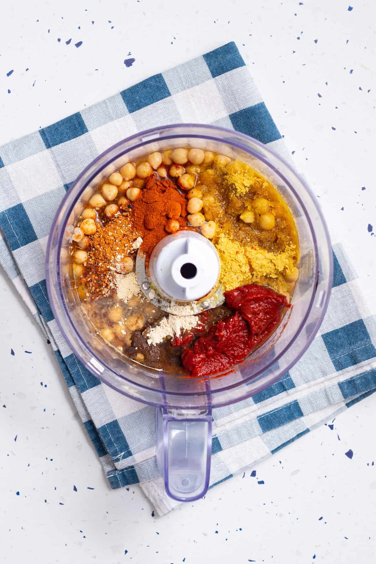 Overhead view of a food processor with chickpeas, no-beef broth, olive oil, nutritional yeast, tomato paste, liquid smoke, maple syrup, smoked paprika, pork seasoning, ground cloves, onion powder, and garlic powder.