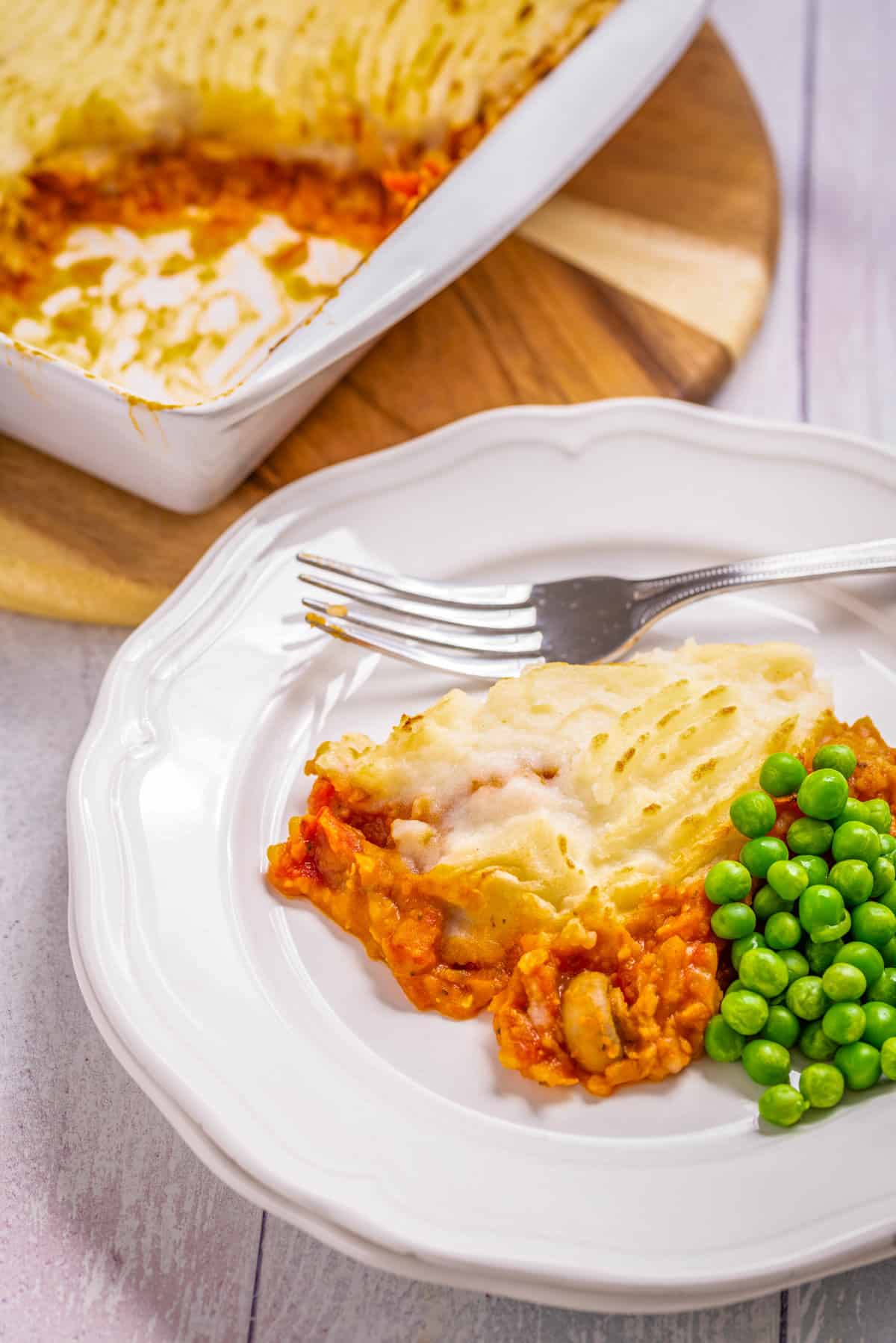 Overhead close-up view of a portion of vegan shepherd's pie with peas and fork on the sides placed on a white plate.
