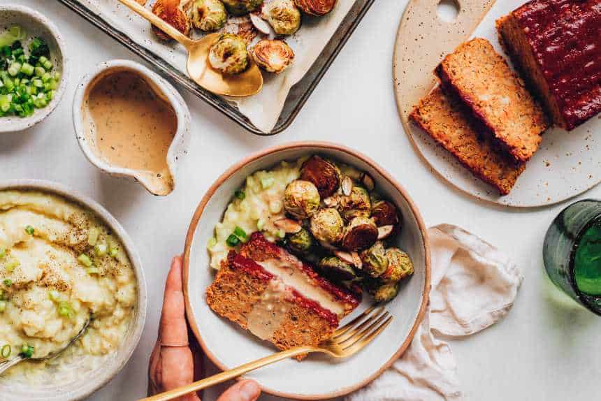 An overhead view of a vegan Thanksgiving dinner bowl with a loaf, mashed potatoes, and roasted Brussels on the side. 