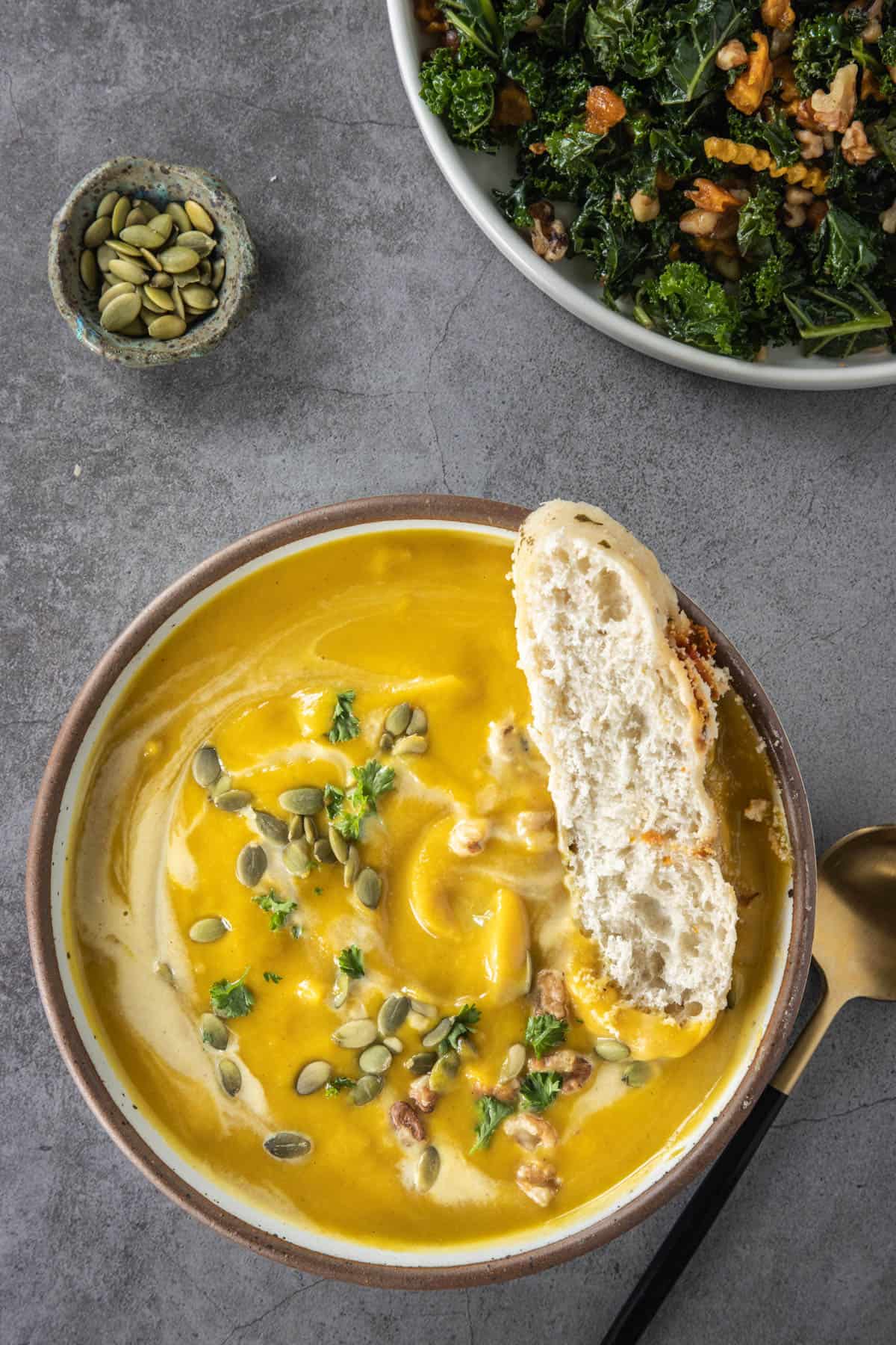 Bowl of pumpkin soup with bread in it, garnish pumpkin seeds and salad in the background