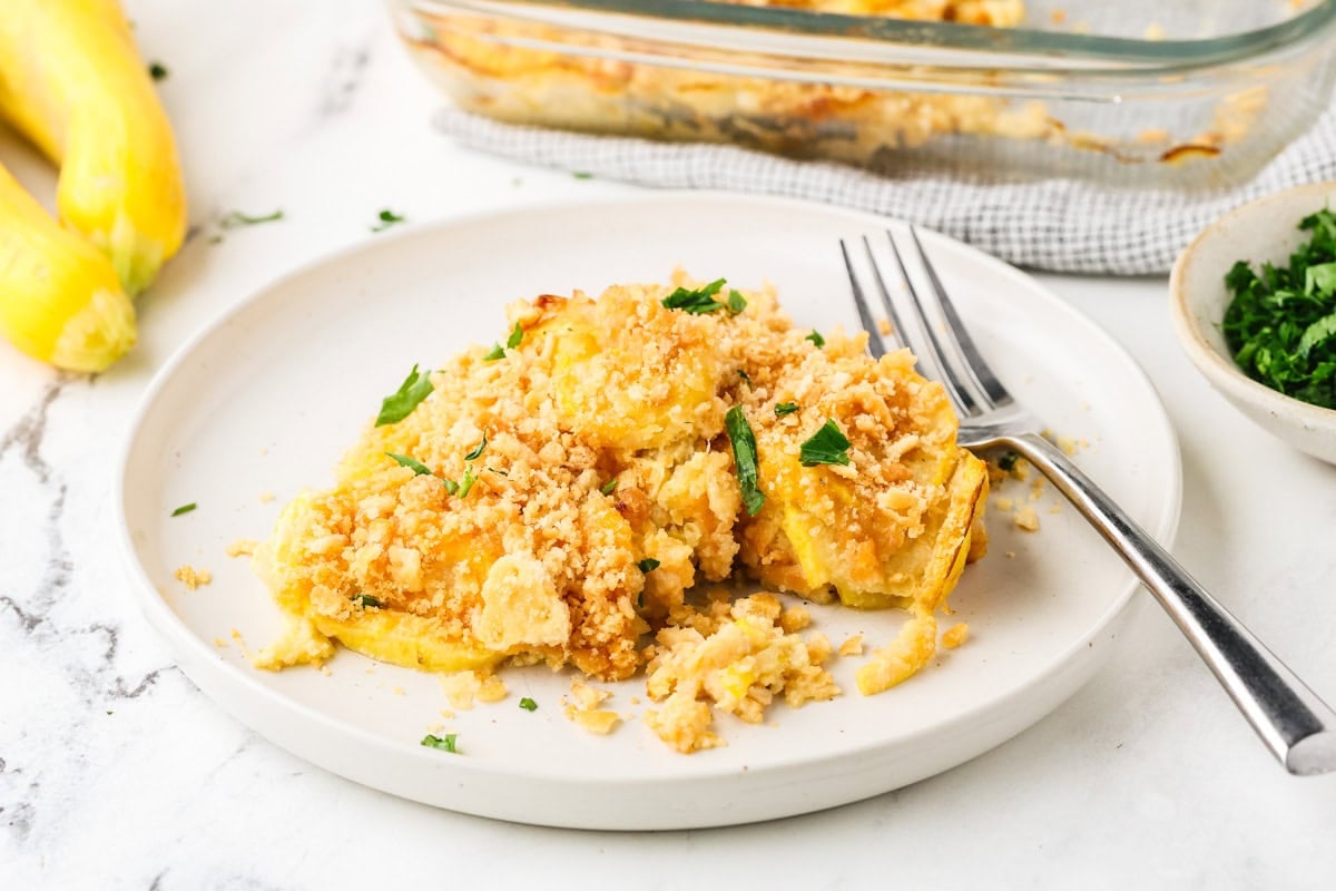 A serving of yellow squash casserole with a crispy cracker topping plated alongside the baking dish.