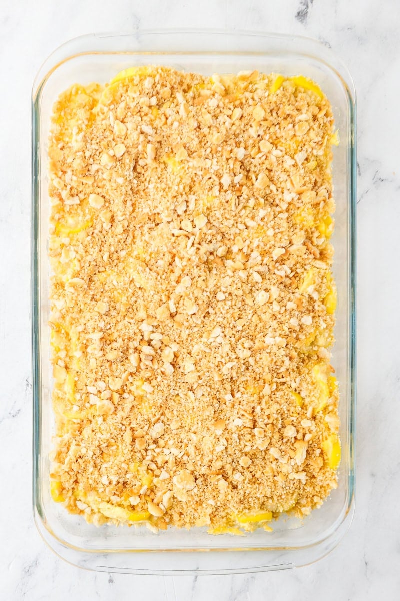 Unbaked yellow squash casserole in a baking dish topped with shredded cheese and crushed buttery crackers.