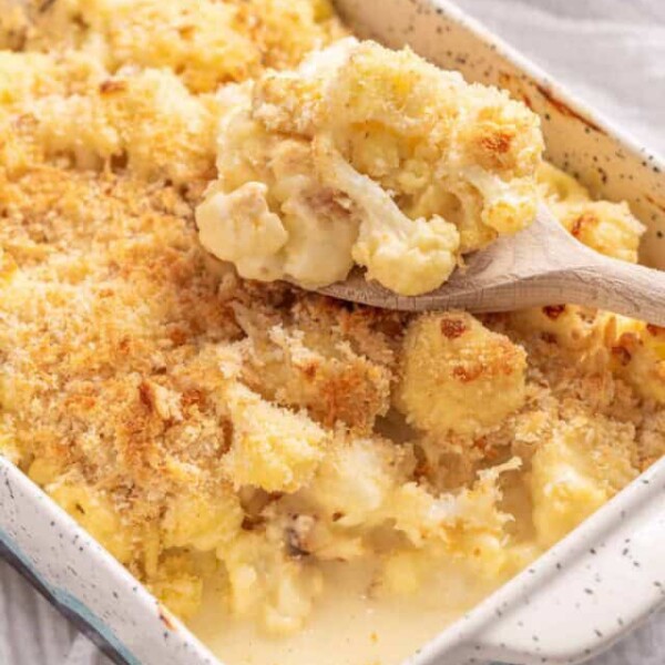 An image of baked cauliflower with cheese in a baking dish, with a full serving spoon resting on top of it,