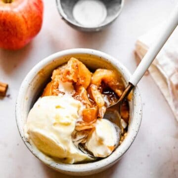 Overhead view of a serving of Bisquick apple cobbler in a white ramekin with a spoon and vanilla ice cream on top.