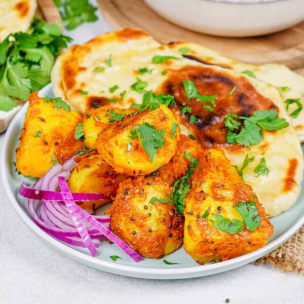 Horizontal shot of Bombay Potatoes on a plate with naan, with extra naan and red onions on the side.