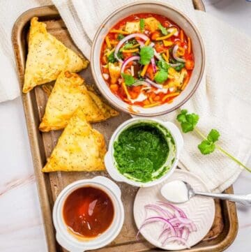 Overhead view of a metal tray with a bowl of samosa chaat and small plates of cilantro chutney, red onions, sweet tamarind chutney, and three pieces of samosas.
