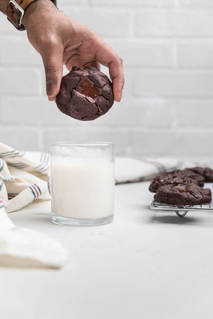 A straight view of a person holding a dairy-free triple chocolate chip cookie to be dipped in a glass of milk and several cookies in the background.