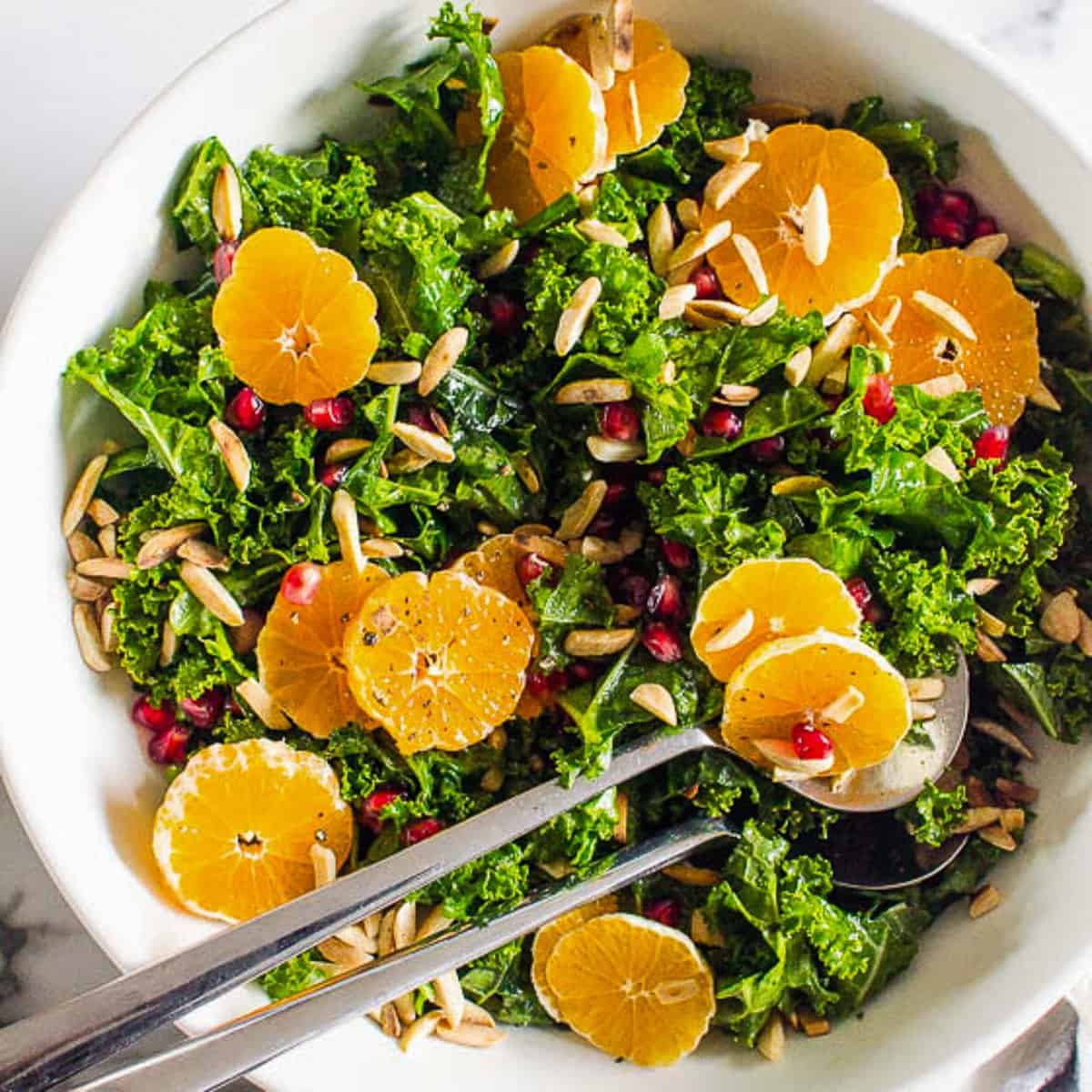 Overhead view of a massaged kale salad with orange-ginger vinaigrette in a white bowl with two serving spoons in it.