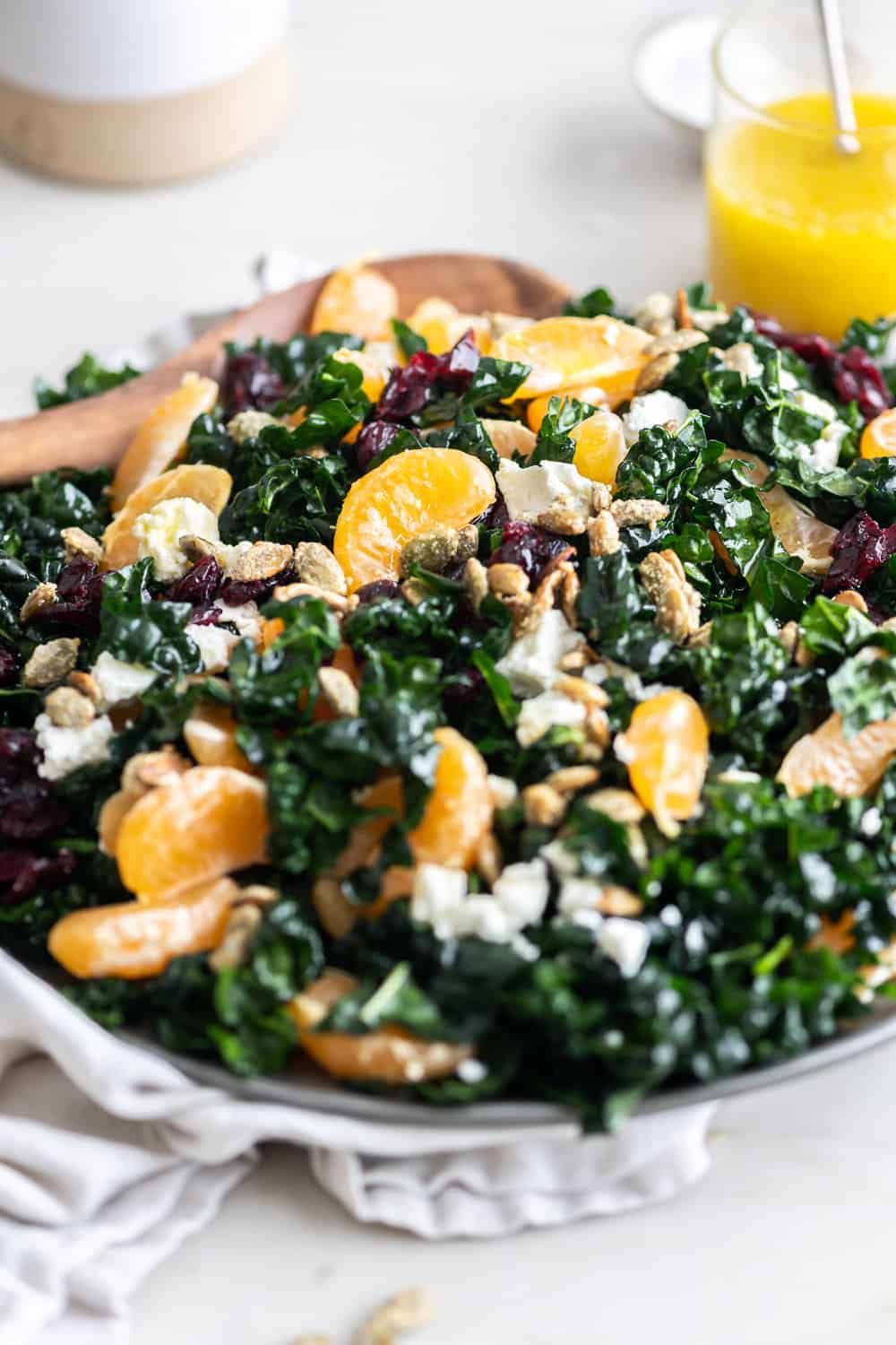 Close-up view of a Tuscan kale salad on a white plate topped with mandarins, cranberries, goat cheese, and candied pepitas.
