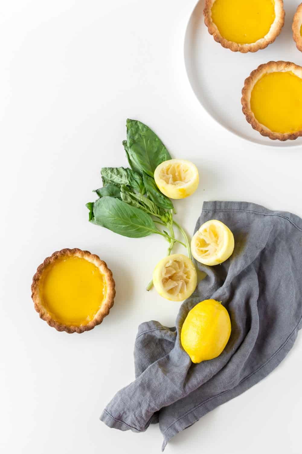 Overhead view of lemon custard tart against a white marble backdrop, with basil leaves, lemons and a grey napkin.
