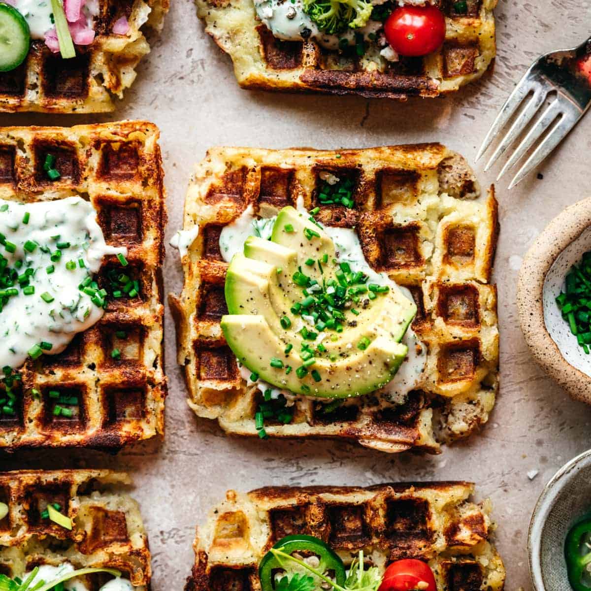 Overhead view of mashed potato waffles on a sand-colored tabletop, with avocado slides on top as garnish.