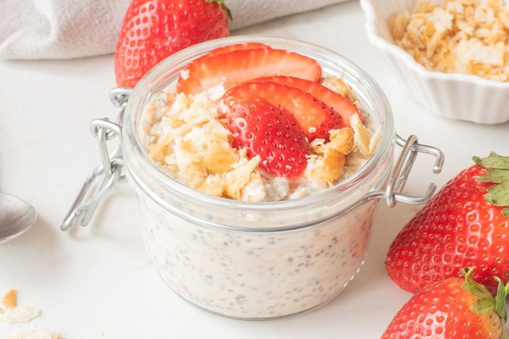 Strawberry cheesecake overnight oats in a glass jar topped with sliced strawberries and graham cracker crumbs.