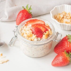 Strawberry cheesecake overnight oats in a glass jar topped with sliced strawberries and graham cracker crumbs.