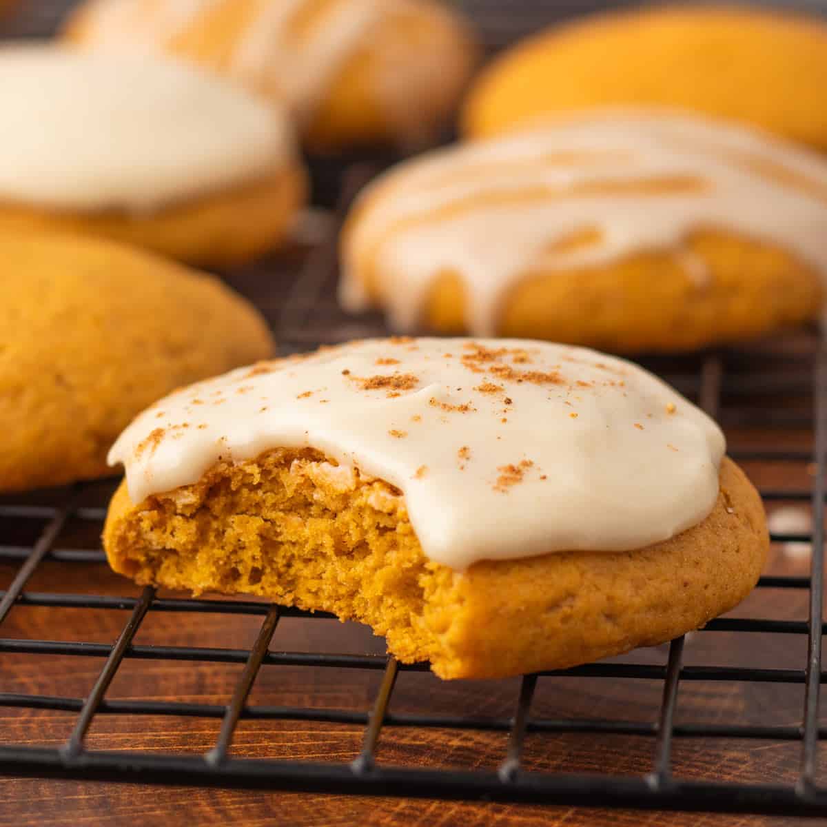 A close up shot of vegan pumpkin cookies with dairy-free cream cheese on top, one with a bite on the front and placed on a cooling rack.