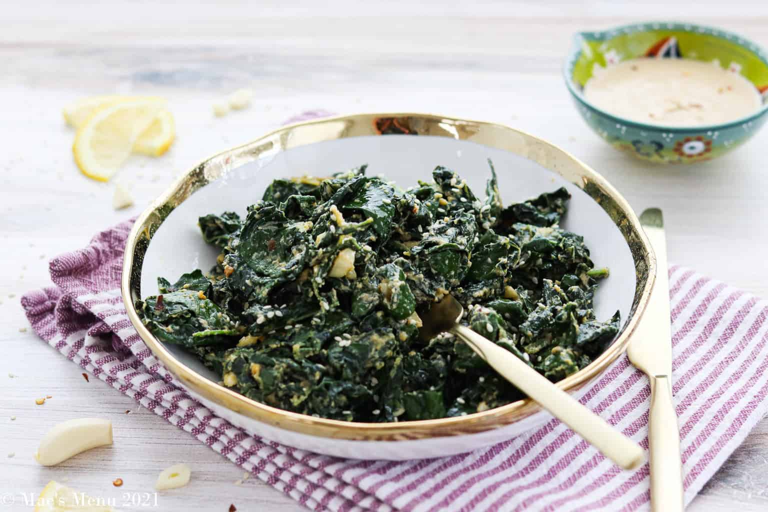 A straight view of a plate of warm kale salad with lemon tahini dressing with a serving spoon above a stripe-colored tablecloth.